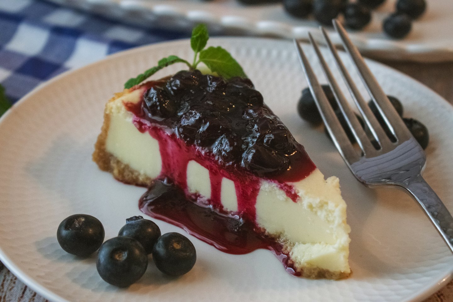 Cheesecake slice with blueberry topping and mint on a ceramic plate.